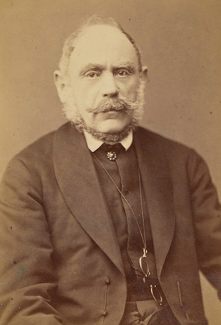 Zdjęcie przedstawia starszego mężczyznę. Jest łysiejący, ma bujne wąsy oraz bokobrody. Ubrany jest w surdut.