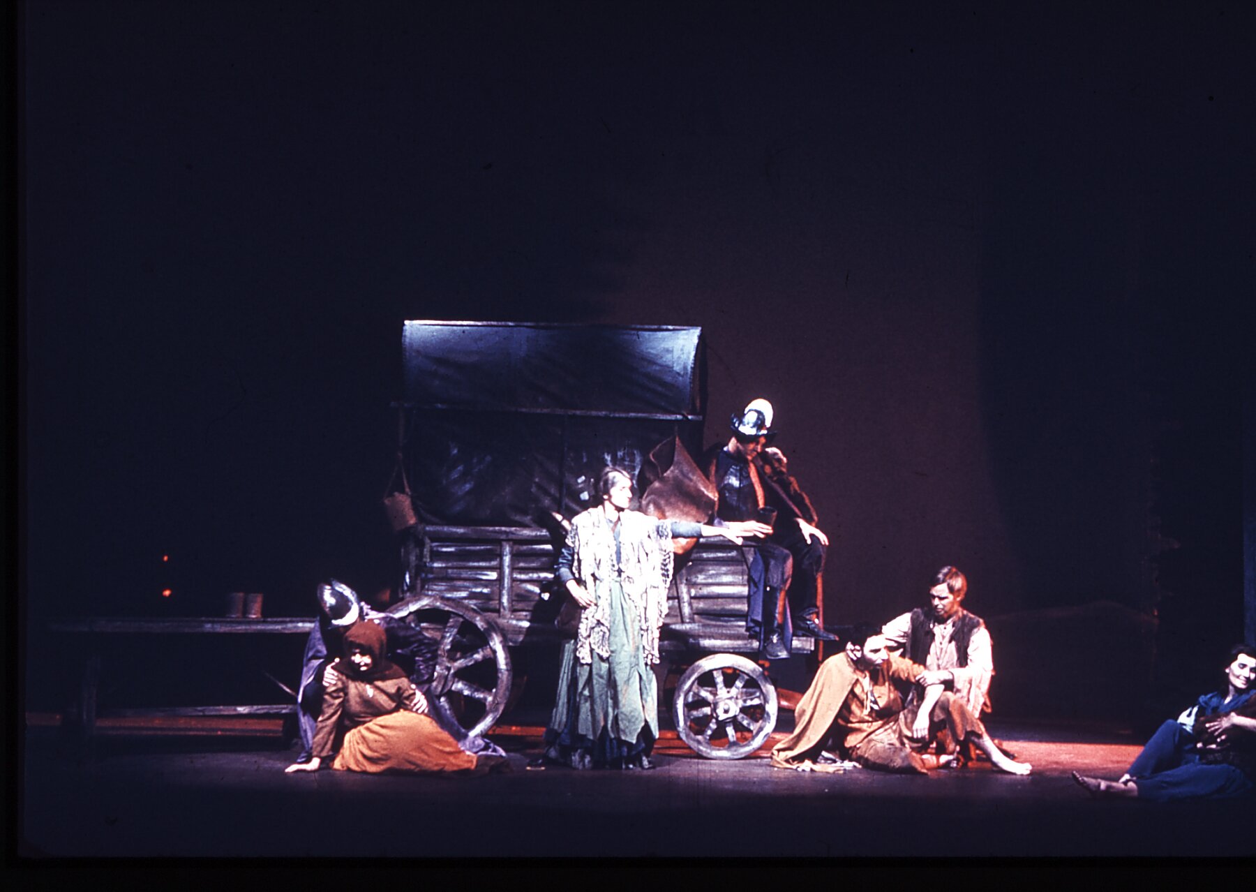 Ilustracja przedstawia grupę osób znajdującą się na scenie teatralnej, przy drewnianym wozie. Wóz ma drewniane koła, tylne o większej średnicy niż przednie. Od góry ma zamontowaną na półokrągłym stelażu materiałową osłonę. Do wozu nie jest zaprzęgnięte żadne zwierzę. Z przodu wozu, w miejscu woźnicy siedzi mężczyzna w stalowym hełmie, butach z wysokimi cholewami i płaszczu. Dłonie ma oparte ma kolanach, głowę spuszczoną, patrzy w ziemie. Przed wozem, w centralnym punkcie stoi kobieta ubrana w długą, skromną suknię, na ramionach ma narzucony długi i szeroki szal z materiału. Lewą rękę kobieta ma podniesioną na bok, do góry. Wskazuje nią na osoby siedzące na ziemi po jej lewej stronie. Są to mężczyzna w łachmanach, z bosymi stopami, przy którym siedzi drugi, ubrany w obszarpaną koszule i kamizelę. Z prawej strony widać jeszcze postać kobiety z bosymi nogami, która siedzi opierając się o osobę, której nie widać z kadrze prezentowanej ilustracji. Po lewej stronie kolejna para, siedząca kobieta koszuli, spódnicy i w chustce a głowie. Za kobietą kuca mężczyzna w metalowym hełmie, obejmuje kobietę za ramiona.