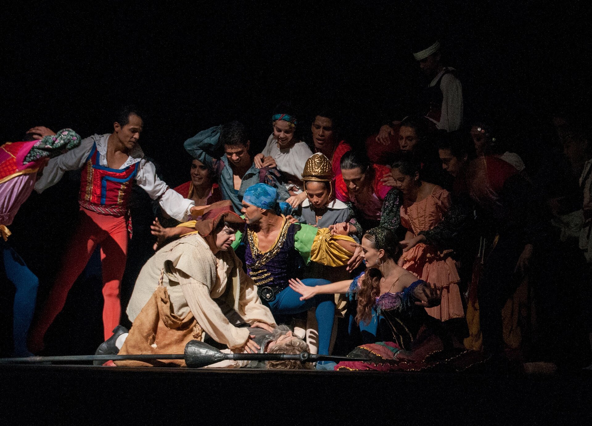 Zdjęcie przedstawia scenę, na której widoczni są aktorzy w trakcie spektaklu. Widoczna jest duża grupa osób ubranych w kolorowe kostiumy, którzy stoją, pochylają się i klęczą wokół postaci w zbroi leżącej na podłodze.