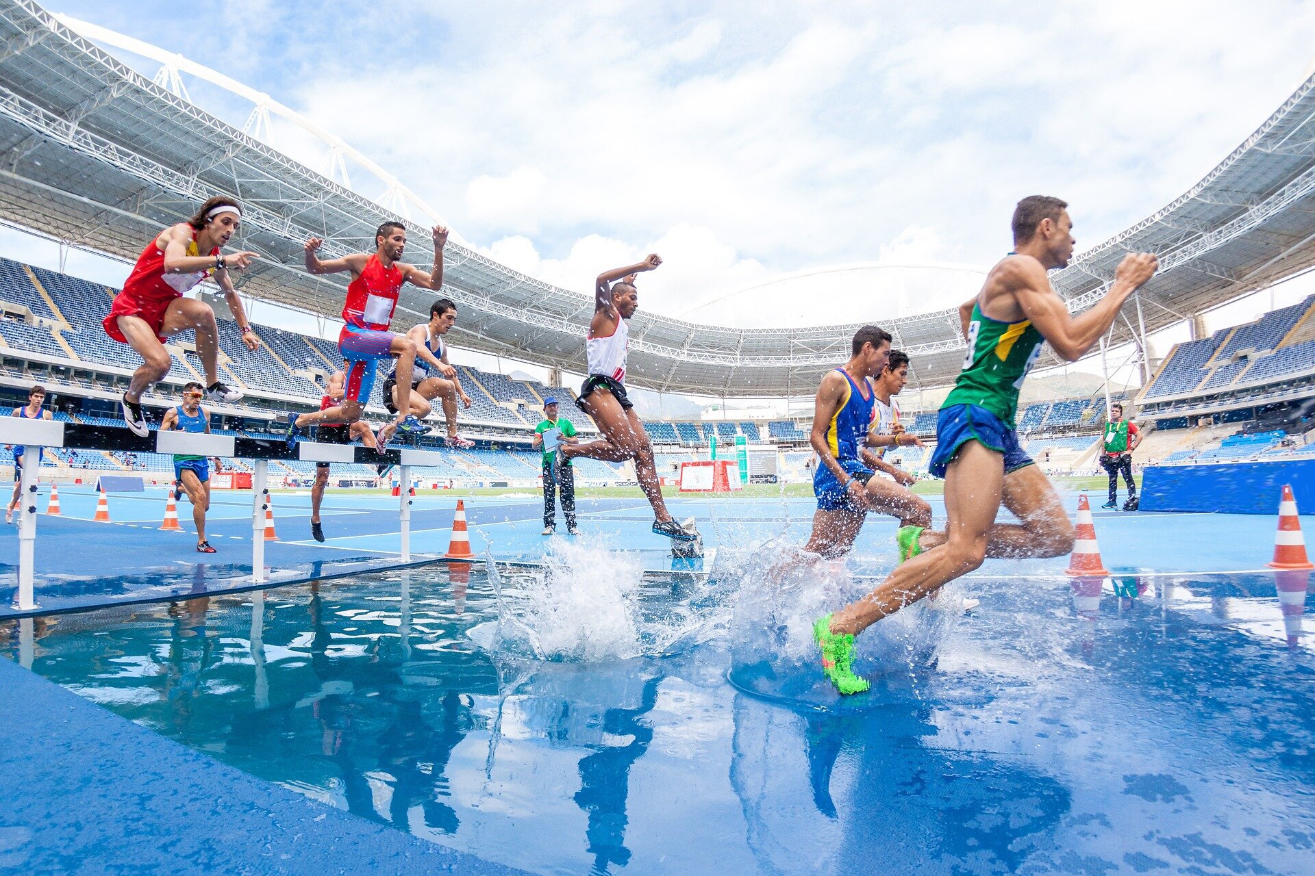 Zdjęcie przedstawia grupę mężczyzn w sportowych strojach biegnących przez wypełniony wodą, płytki basen umieszczony na stadionie sportowym.