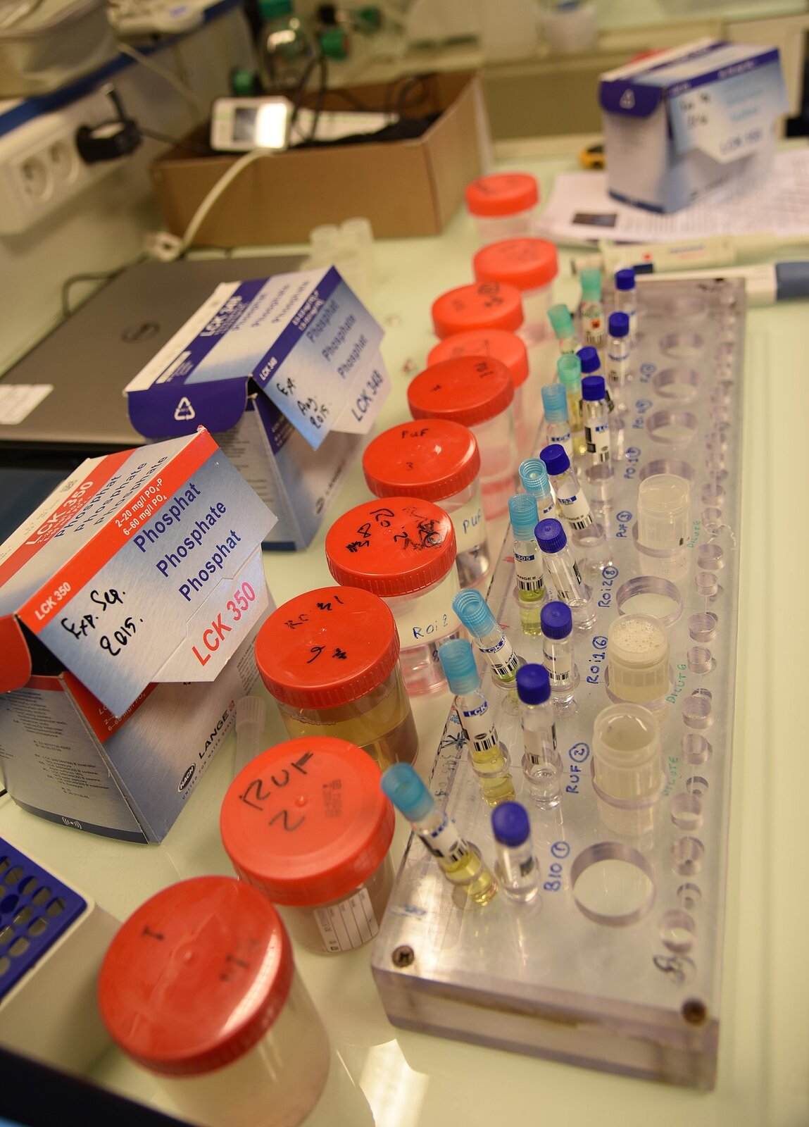 Fotografia przedstawiająca kilkadziesiąt próbek wody w plastikowych lub małych szklanych pojemnikach.