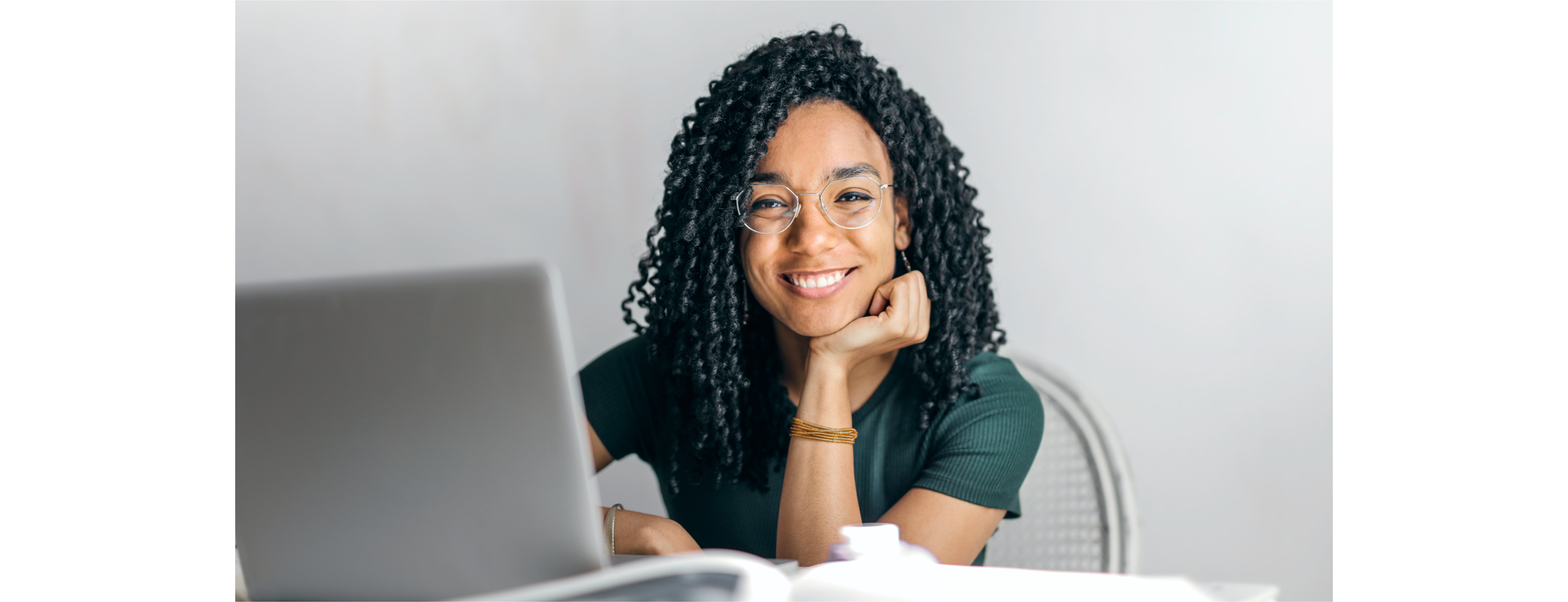 Zdjęcie przedstawia młodą, uśmiechniętą dziewczynę w okularach z kręconymi włosami. Dziewczyna siedzi przodem do kadru przed otwartym laptopem. 