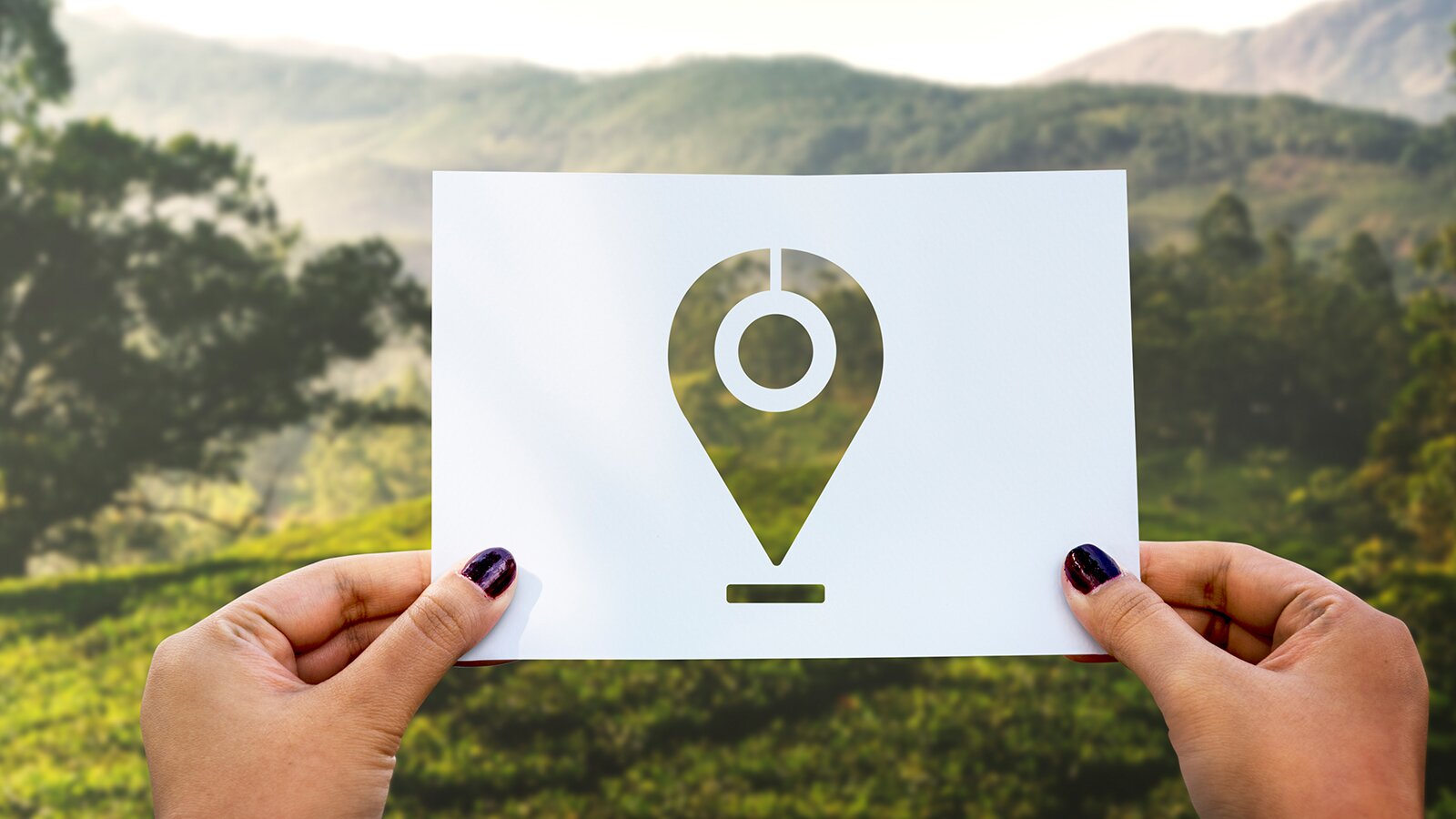 Zdjęcie przedstawia dłonie, które trzymają kartkę z wyciętym znakiem lokalizacyjnym. W tle widoczny górzysty krajobraz. 