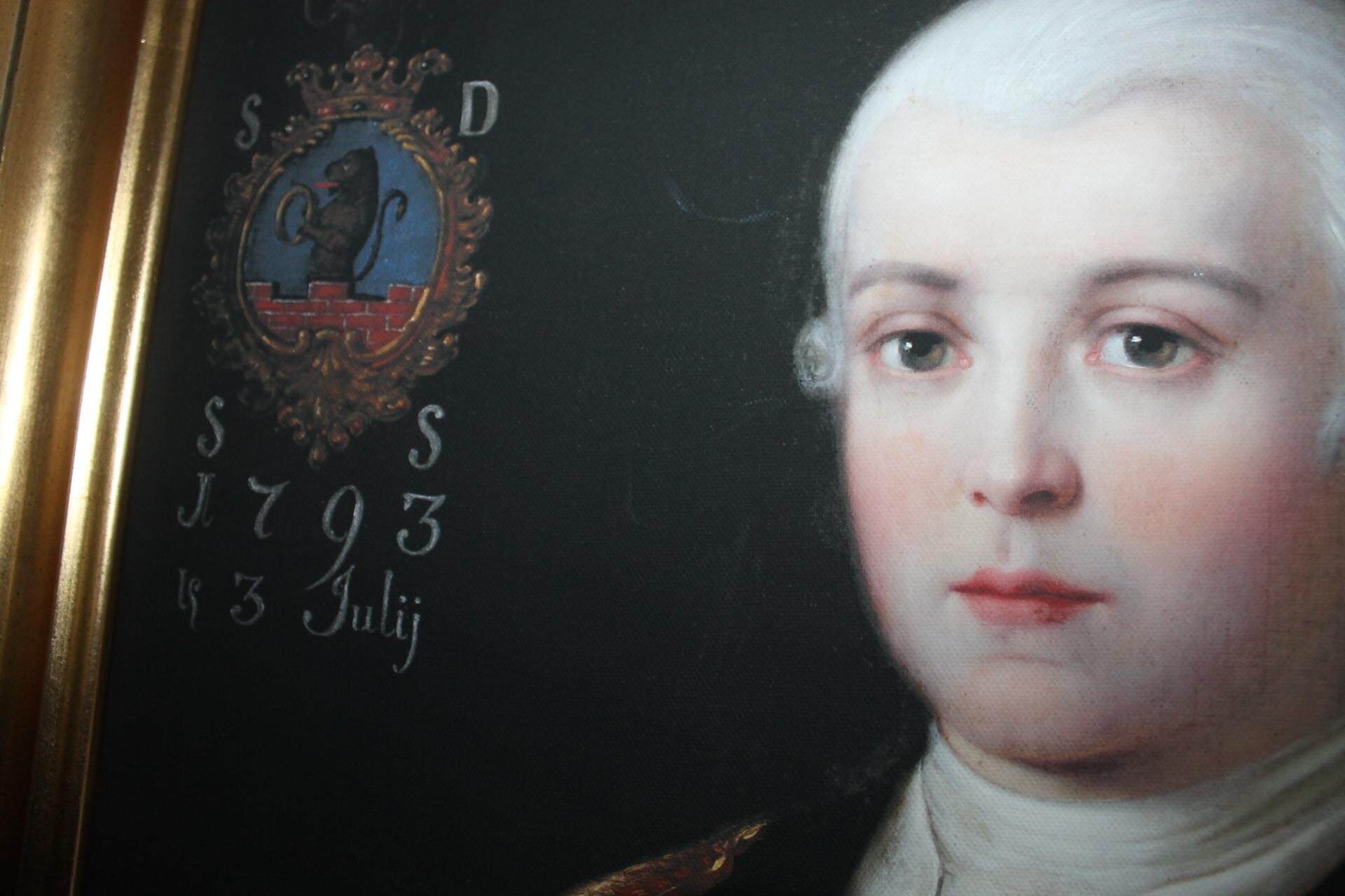 Zdjęcie przedstawia portret mężczyzny z białymi włosami i zielonymi oczami. Ukazane jest zbliżenie na samą twarz. Mężczyzna ma obojętny wyraz twarzy. Za jego plecami na ciemnej ścianie namalowany jest herb szlachecki - ceglany mur ze stojącym na nim gryfem trzymającym elipsę. Wokół herbu namalowane są białe litery i cyfry.