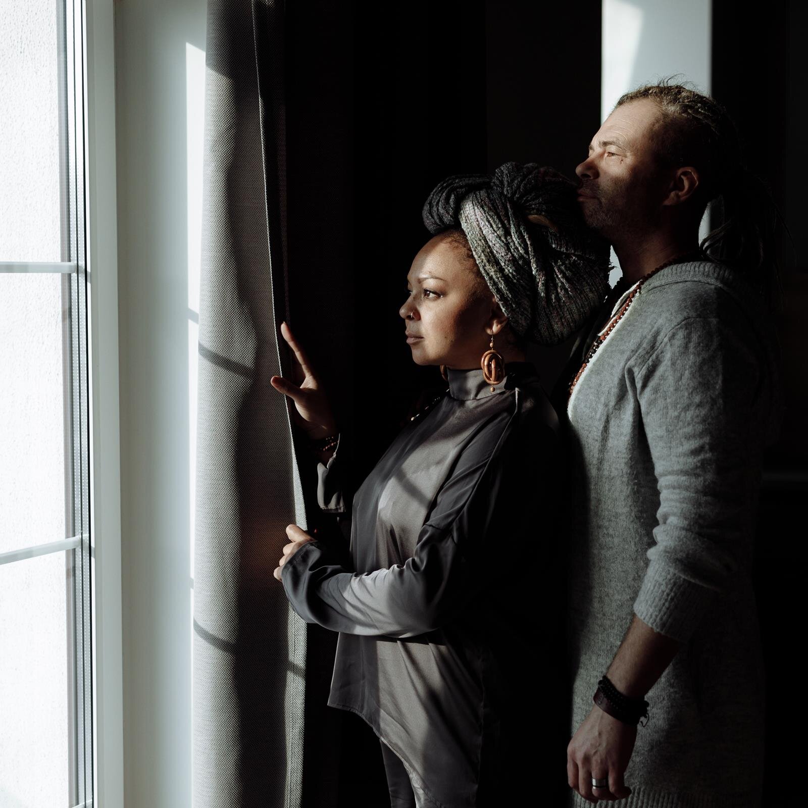 Zdjęcie przedstawia parę stojącą bokiem do patrzącego. Kobieta po lewej stronie odsłania zasłonę i patrzy przez okno. Za nią stoi mężczyzna w szarej koszulce z długimi rękawami, również spoglądający przez okno.