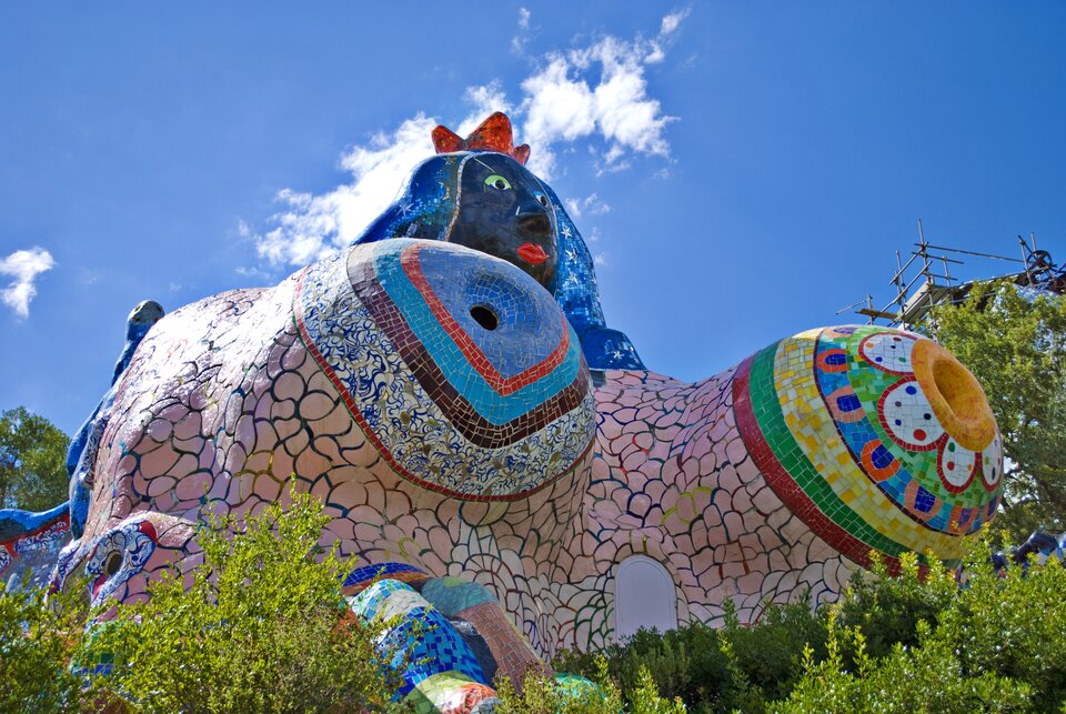 lustracja przedstawia pracę Niki de Saint Phalle. Jest to budynek‑rzeźba Cesarzowa. Fotografia została wykonana w 2010 roku. Budowlę możemy oglądać w Capalbio w Toskanii we Włoszech. Na zdjęciu widzimy kolorową rzeźbę przypominającą kobietę z dużymi piersiami. Twarz oraz głowa kobiety są w kolorze niebieskim. Na głowie bohaterka ma czerwoną koronę. Dookoła widoczny jest park. Autorką pracy jest francuska rzeźbiarka oraz malarka. W swoich pracach bardzo często wykorzystywała motyw kobiecego ciała.