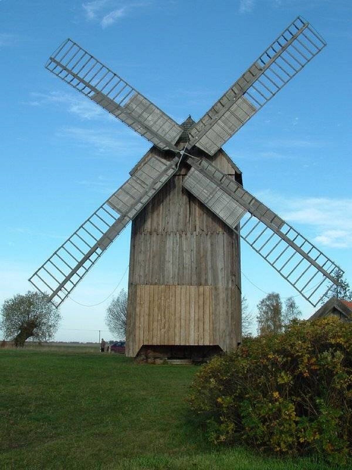 Zdjęcie ukazuje drewniany, brązowy wiatrak. Wiatrak ma cztery skrzydła, poruszające się podczas wiatru. W oddali rosną drzewa. W tle rozciąga się błękitne niebo.