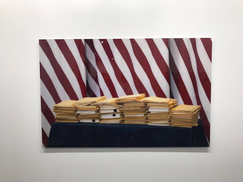 Ilustracja przedstawia fotografię Thomasa Demanda pt. „Folders”. Ukazuje ona stosy jasnobrązowych teczek z dokumentami w środku, które ułożone są na stole. W tle widoczna jest kotara w biało-czerwone, ukośne paski.