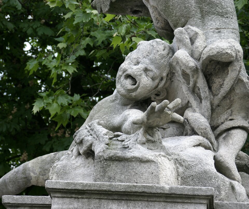 Zdjęcie przedstawia fragment jasnej rzeźby dziecka wyciągającego przed siebie dłoń w geście rozpaczy. Rzeźba wykonana jest z jasnego materiału. Rzeźba jest zniszczona, na przykład na twarzy dziecka brakuje nosa.  