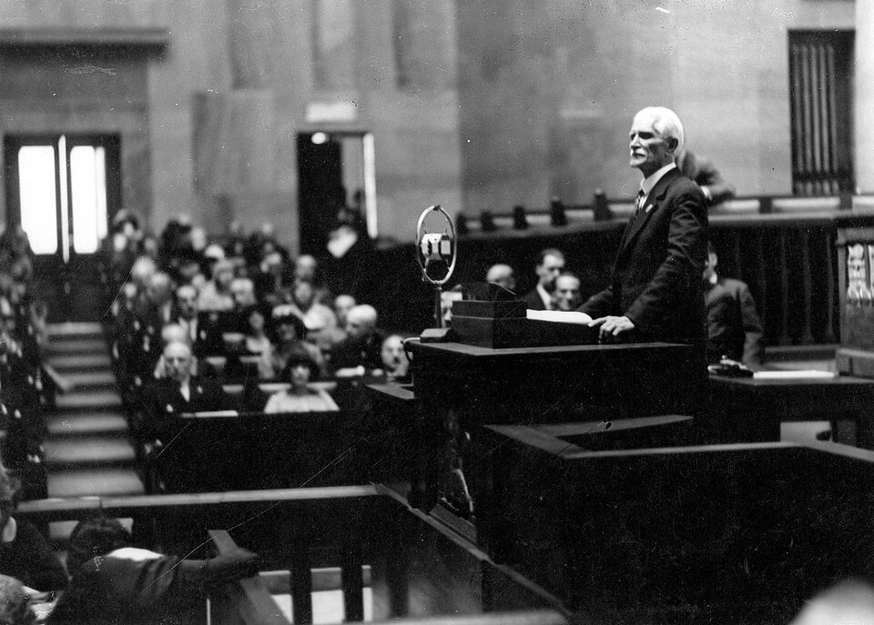 Ilustracja przedstawia mężczyznę ubranego w garnitur i koszulę. Ma on siwe włosy i krótką brodę. Przemawia na mównicy w sejmie. Na trybunach siedzą ludzie. 