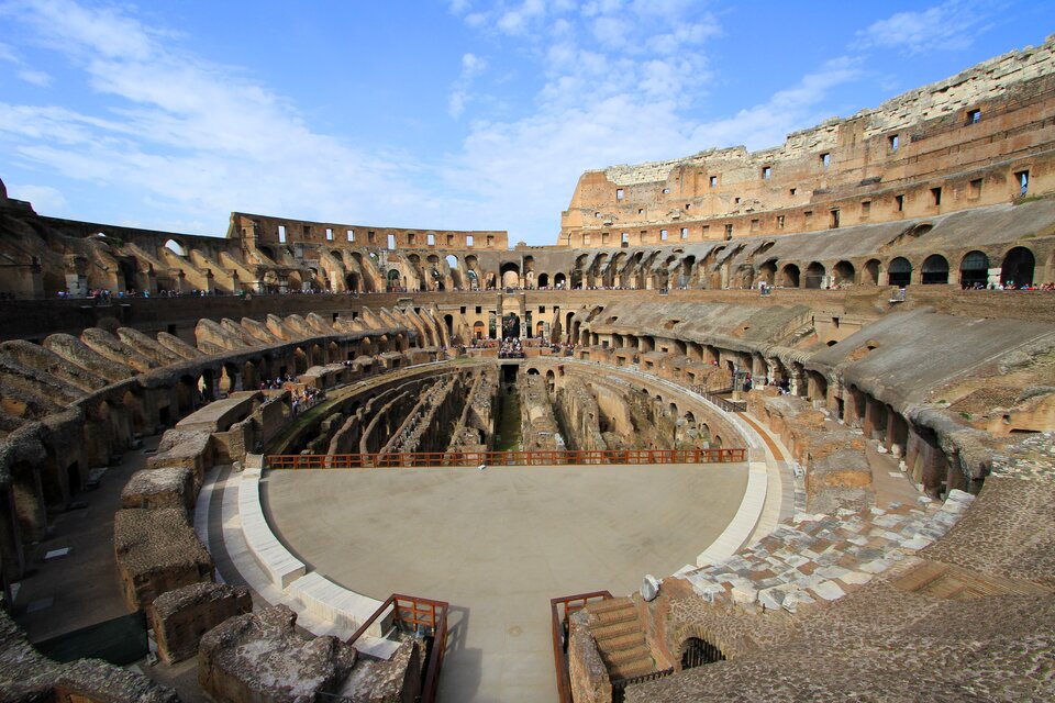 Fotografia przedstawia Koloseum widoczne od wewnątrz. Na pierwszym planie - scena ogrodzona współczesnymi barierkami. Na bocznych częściach sceny znajdują się trybuny umiejscowione w prawym i lewym łuku, a w przedniej jej części jest wejście oraz kilka betonowych schodków. Wokół konstrukcji umieszczono kilka pięter trybun, kolumny połączone od góry łukiem oraz prostokątne otwory okienne. W oddali przechadzają się turyści. Zdjęcie zostało zrobione w ciągu dnia, o lekko pochmurnym niebie.