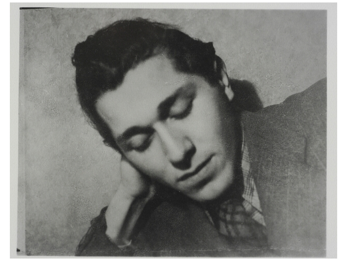 Zdjęcie przedstawia młodego mężczyznę, który opiera głowę na prawej dłoni. Mężczyzna ma pociągłą twarz, wyraziste, ciemne brwi i wydatny nos i usta. Ma zamknięte oczy. Jego włosy są ciemne i krótkie, zaczesane ku górze. Mężczyzna jest w koszuli i garniturze.