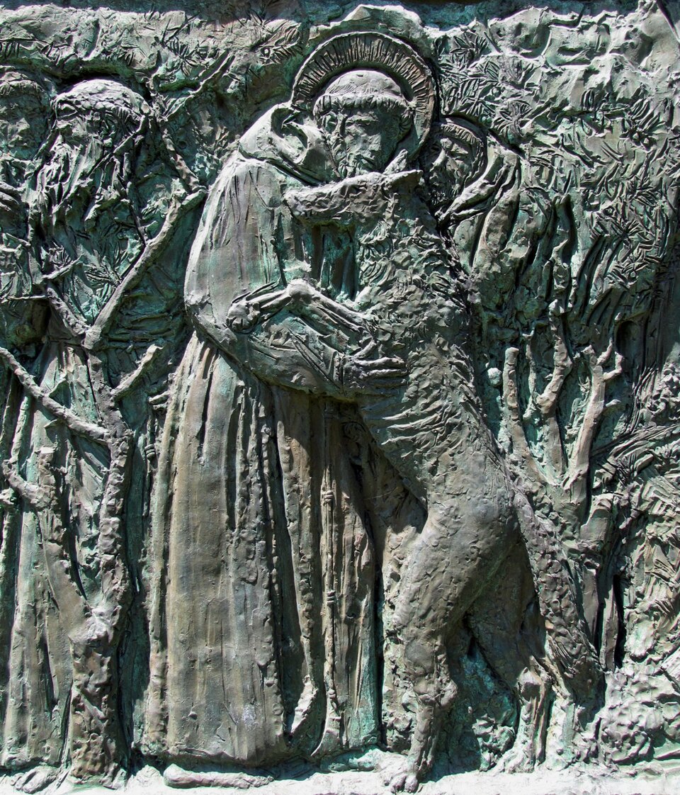 Płaskorzeźba przedstawia mężczyznę, który przytula stojącego na dwóch łapach wilka. Mężczyzna nosi habit z kapturem. Ma on brodę, a na głowie wygoloną tonsurę (wygolone włosy na czubku głowy zakonników miały przypominać koronę cierniową). Głowę okala aureola. Stojące obok mężczyzny zwierzę sięga mu do brody. Wilk ukazany został z lewego profilu i patrzy na wprost. W tle znajdują się drzewa, krzewy i postaci ludzi. 