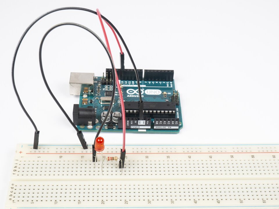 Zdjęcie przedstawia płytkę Arduino oraz sześćdziesięciowierszową płytkę stykową. Wiersze podpisano co pięć zaczynając od 1. Po lewej stronie podpisano poszczególne kolumny. Na górnym obszarze: j i h g f. Na dolnym obszarze: e d c b a. Na płytce stykowej znajduje się czerwona dioda LED, rezystor oraz czarny przewód. Ze złącza 13 Arduino poprowadzono czerwony przewód, którego koniec umieszczono na płytce stykowej w kolumnie h w wierszu 24. Ze złącza GND Arduino poprowadzono czarny przewód, którego drugi koniec umieszczone na płytce stykowej na szynie minus. Pierwszy koniec kolejnego czarnego przewodu umieszczono w kolumnie j w wierszu 18, a drugi koniec w kolumnie h w wierszu 24. Pierwszą nóżkę diody LED umieszczono w kolumnie h w wierszu 18, a drugą w kolumnie h w wierszu 20. Pierwszą nóżkę rezystora umieszczono w kolumnie f w wierszu 20, a drugą w kolumnie f w wierszu 24.