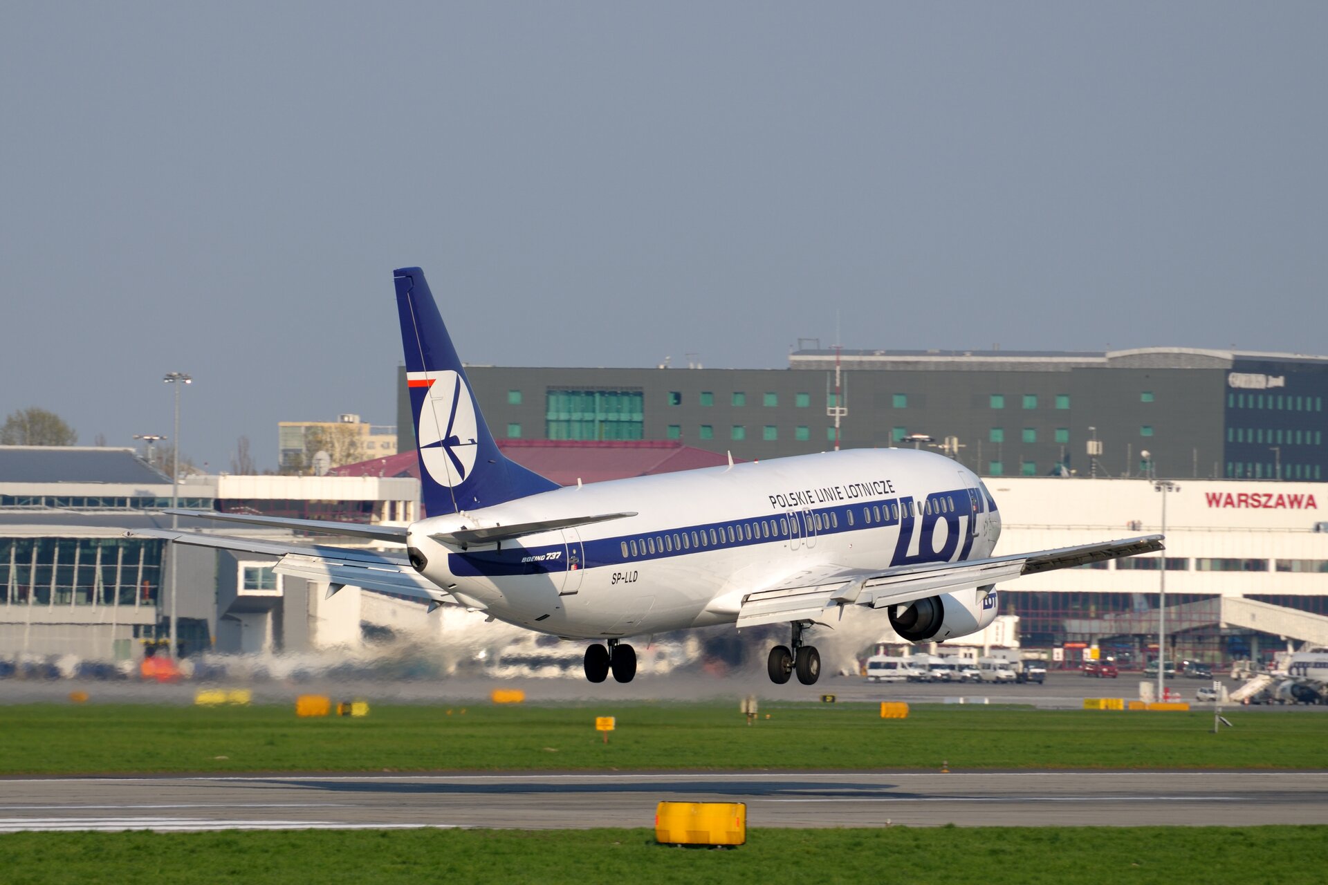 Fotografia ukazuje duży pasażerski samolot startujący do lotu. W tle budynki portu lotniczego.