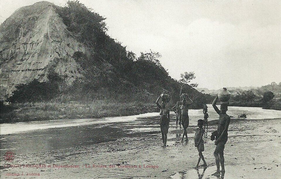 Zdjęcie przedstawia grupę półnagich, czarnoskórych kobiet i dzieci, które znajdują się przy rzece. Kobiety niosą na głowach dzbany z wodą. Za nimi w tle jest wysokie wzniesienie oraz lasy.