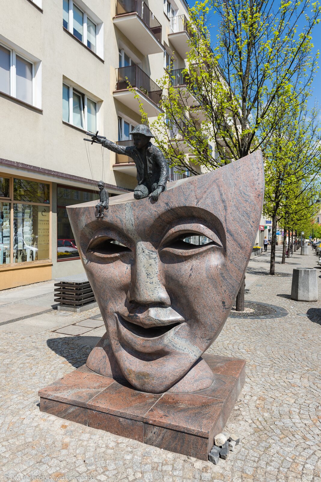 Zdjęcie przedstawia rzeźbę autorstwa Bogusława Szycika pod tytułem „Maska”. Na zdjęciu znajduje się rzeźba przedstawiająca maskę z odciętą górną częścią. Maska wyraża emocje szczęścia. Na głowie maski siedzi chłopiec w czapce w ręku trzymający kukiełkę na sznurkach. Rzeźba jest wykonana z brązowego kamienia. W tle zdjęcia znajduje się drzewo oraz budynki.
