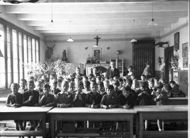 Fotografia przedstawia klasę w jednej ze szkół. Klasa jest bardzo liczna. Uczniowie siedzą w ławkach i patrzą w obiektyw. Z tyłu klasy widać czworo nauczycieli, trzy kobiety i mężczyznę. Na ścianie wisi krzyż z Chrystusem poniżej obraz z postacią w mundurze. W rogu wisi wypchana głowa jelenia.
