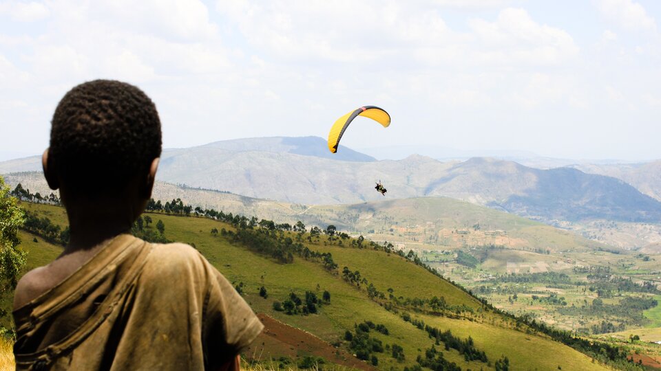 Zdjęcie przedstawia Burundi. Na pierwszym planie tyłem stoi czarnoskóry chłopiec. Za nim są zbocza porośnięte trawą i drzewami. Nad nimi lata paralotniarz. W tle są góry.