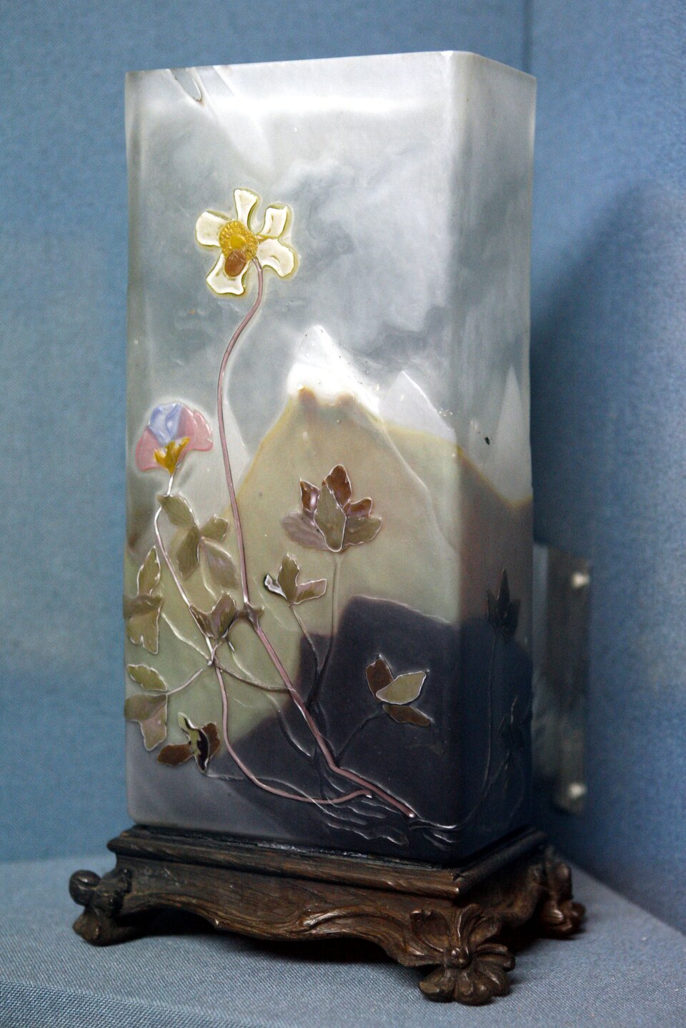 Ilustracja przedstawia wazon autorstwa Émile Gallé. Wazon ozdobiony jest motywami nawiązującymi do natury. Na pierwszym planie jest motyw roślinny, po czym w tle widać krajobraz górski. Wazon ma kształt prostokątny.