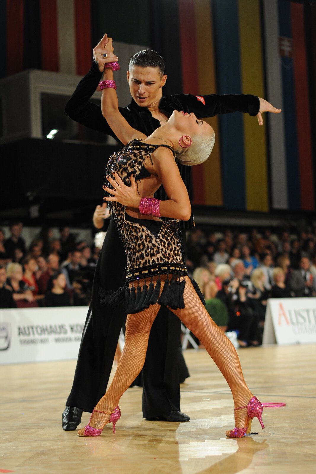 Fotografia przedstawia parę w tańcu. Kobieta ma na sobie sukienkę w panterkę zakończoną czarnymi frędzlami. Na stopach ma różowe sandały na obcasie. Ma duże, różowe, okrągłe kolczyki, a na obu nadgarstkach ma wiele bransoletek w kolorze różowym. Kobieta ma blond włosy zaczesane do tyłu oraz paznokcie u rąk pomalowane na kolor czarny. Jej prawa ręka podniesiona jest do góry, a lewa jest na wysokości talii. Kobieta stoi bokiem, a z jej prawej strony stoi ciemnowłosy mężczyzna. Ma na sobie czarną koszulę, spodnie i zakryte, wiązane buty. Jego prawa dłoń dotyka prawej dłoni kobiety, a lewa ręka wyciągnięta jest w bok. Para znajduje się na jasnym parkiecie. W tle znajduje się widownia. Tło jest rozmyte, niewyraźne. 