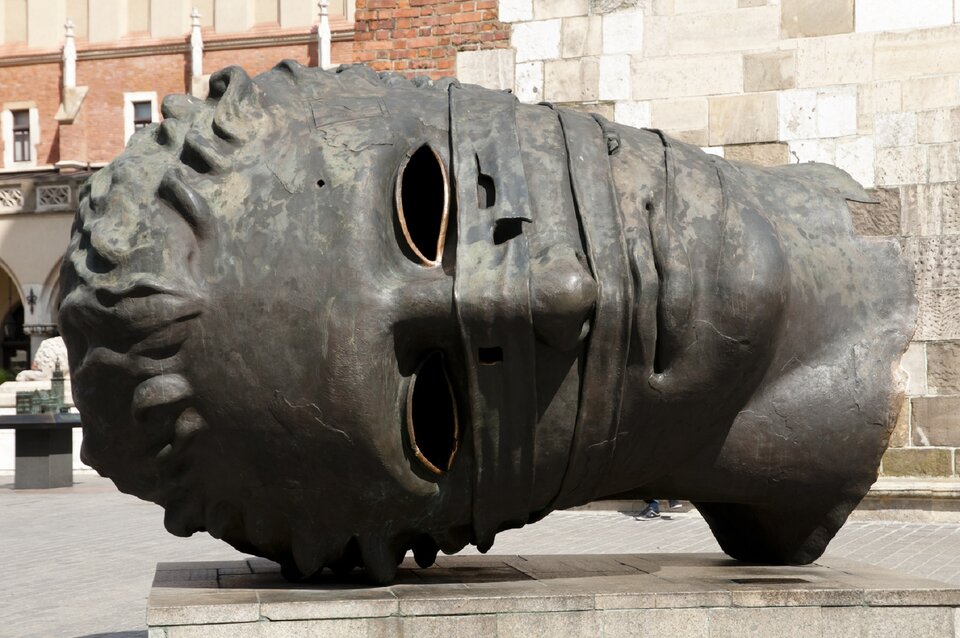 Fotografia przedstawiająca rzeźbę głowy spętanego Erosa wykonaną z brązu. Dużych rozmiarów głowa leży na prawym boku.