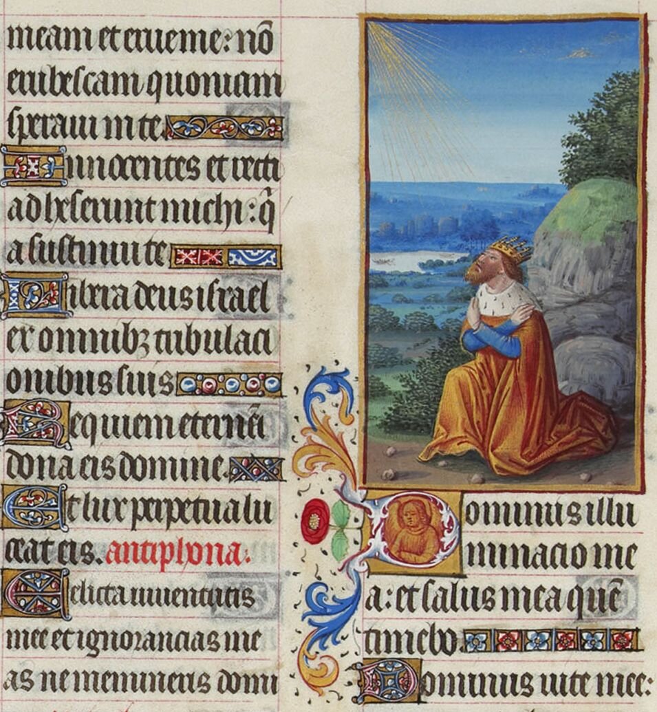 Ilustracja przedstawia bogato zdobioną kartę ze średniowiecznej, ręcznie pisanej książki. Po lewej stronie kartki znajduje się ręcznie pisany tekst. Po prawej stronie ilustracja przedstawia króla modlącego się na wzgórzu. W tle widać drzewa, skały i niewielkie jezioro. Król w koronie na głowie, nosi czerwono‑złoty płaszcz. Klęczy, spoglądając w górę w stronę padających z nieba promieni. Mężczyzna nosi brodę, ręce ma skrzyżowane na piersiach.