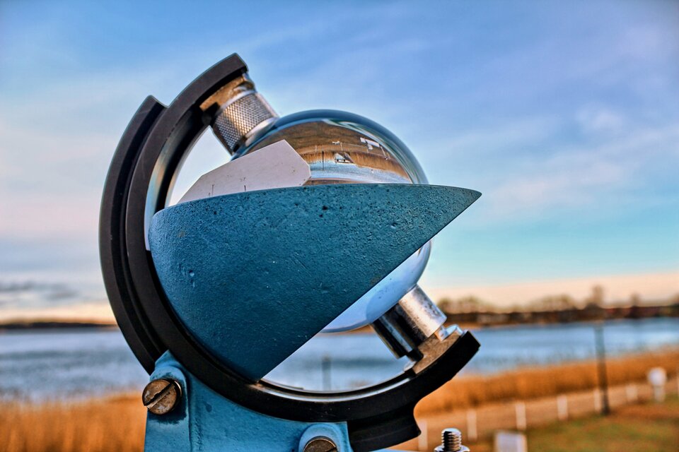 Zdjęcie przedstawia heliograf Campbella‑Stokesa. Składa się on ze szklanej kuli umieszczonej nad metalową kołyską za pomocą półokrągłego uchwytu. W metalowej kołysce znajduje się kawałek papieru heliograficznego. W tle fotografii widać błękitne niebo i powierzchnię wody. 