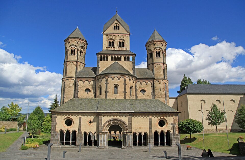 Ilustracja o kształcie poziomego prostokąta przedstawia portal główny kościoła Maria Laach. Budowla składa się z geometrycznych brył w postaci walców i prostopadłościanów. Kościół jest masywny, ma małe okna zakończone półkoliście. Widoczny jest narteks z usytuowaną pośrodku bramą wejściową oraz arkadami po bokach. Za nim symetrycznie rozłożona bryła właściwa: z nawą główną, nawami bocznymi wieżami po bokach. Na środku wznosi się wieża znajdująca się na przecięciu nawy głównej z transeptem.   Elewację zdobi fryz arkadkowy. Po prawej znajduje się fragment innej budowli. Z obu stron kościół otacza pejzaż. Tło wypełnia błękitne niebo z chmurami. 