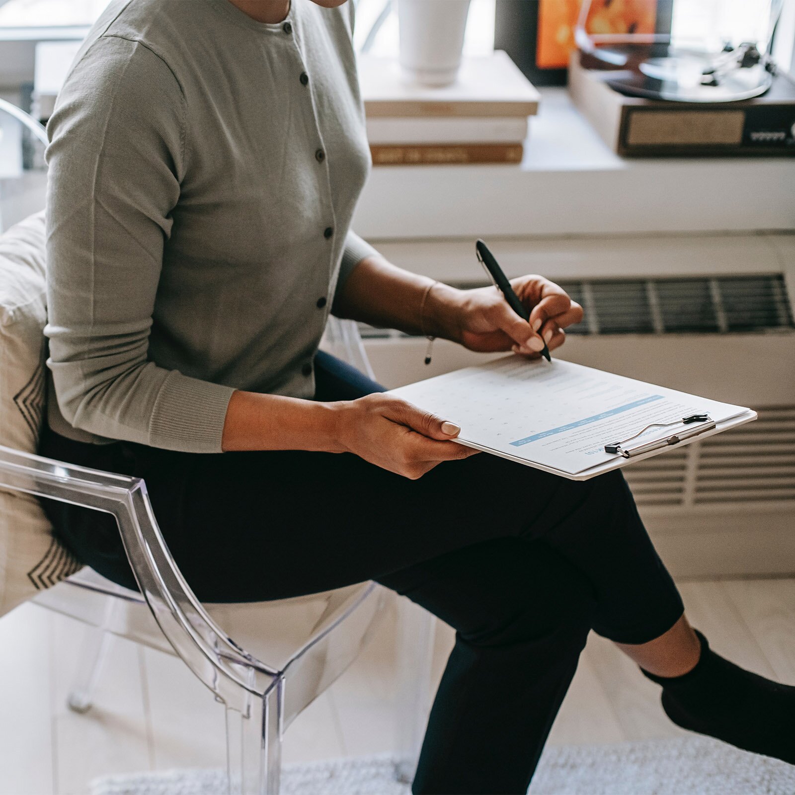 Zdjęcie przedstawia kobietę od szyi w dół, siedzącą na krześle. Postać ma założoną nogę na nogę, na kolanie opiera podkładkę z kartką. Podkładkę podtrzymuje prawą dłonią, a w lewej trzyma długopis.
