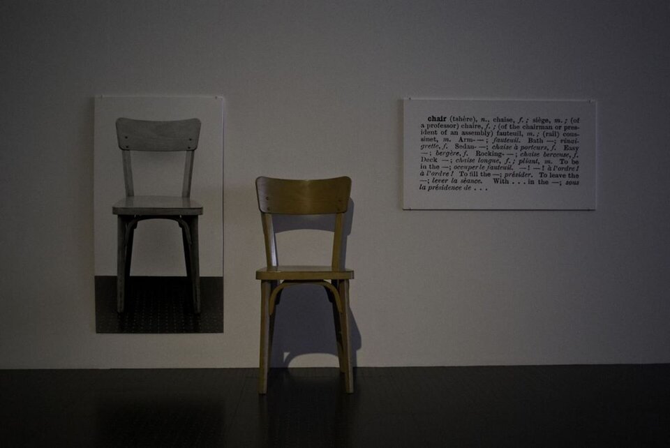 Ilustracja przedstawia instalację Joseph'a Kosuth, „Krzesła Jeden i Trzy". Na zdjęciu znajdują się: krzesło, fotografia krzesła, wyjaśnienie znaczenia słowa "krzesło". Wszystkie elementy są umieszczone obok siebie: krzesło stoi na ciemnej podłodze przy jasnej ścianie, fotografia wisi z lewej strony stojącego krzesła, wyjaśnienie słowa - z prawej strony.