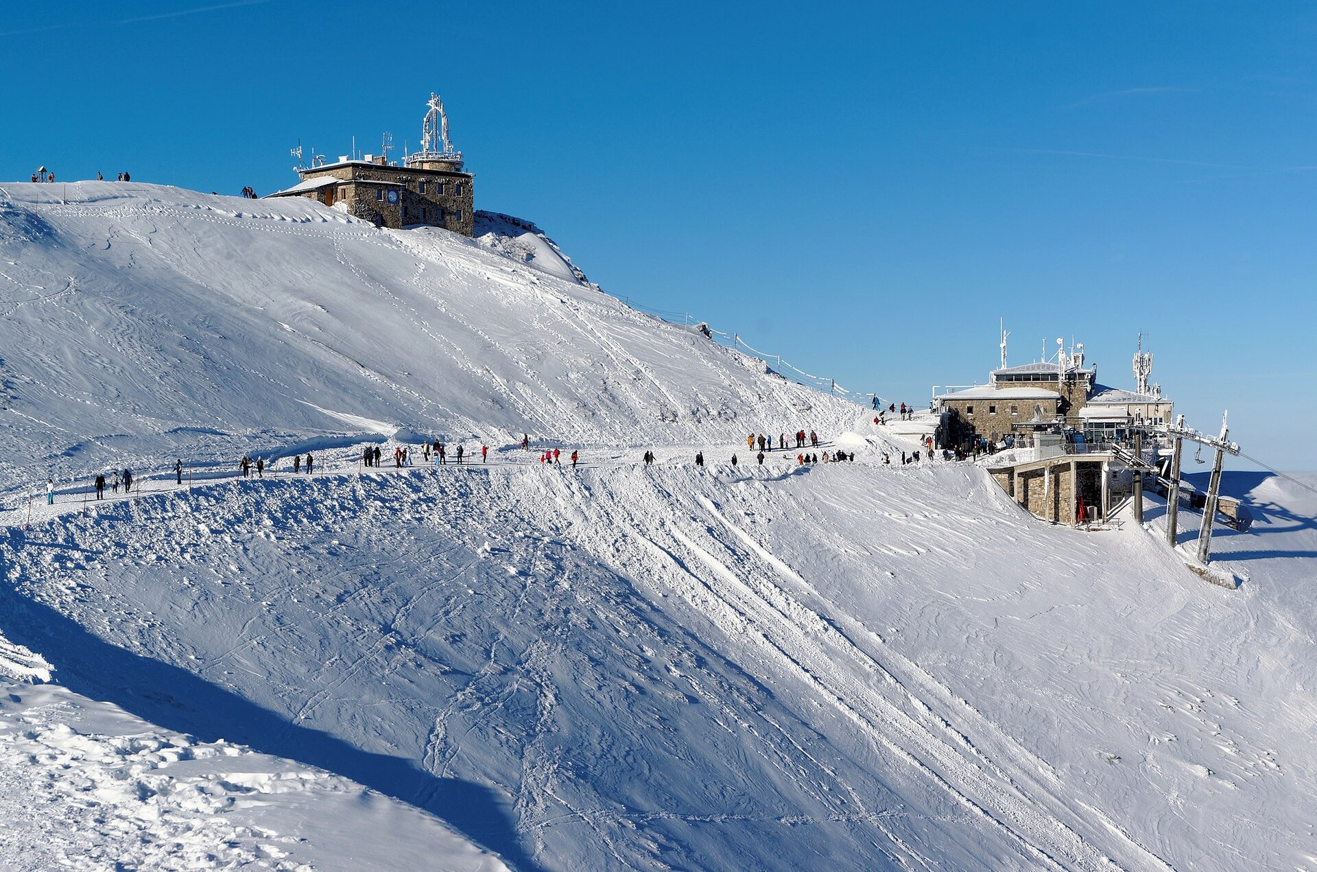 Fotografia przedstawiająca wierzchołek góry w czasie zimy. Teren w całości pokryty śniegiem, na szczycie zabudowania niewielkiej stacji badawczej, poniżej budynek należący do infrastruktury kolejki prowadzącej na górę.