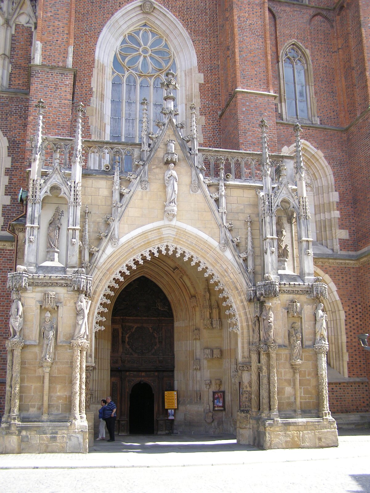 Ilustracja przedstawia wejście główne do katedry św. Jana Chrzciciela we Wrocławiu. Portal ten zawiera ozdobne wieżyczki oraz przedstawienia postaci. Sam portal jest dużo większy od wejścia do świątyni. Przy drzwiach do kościoła widoczni są turyści.