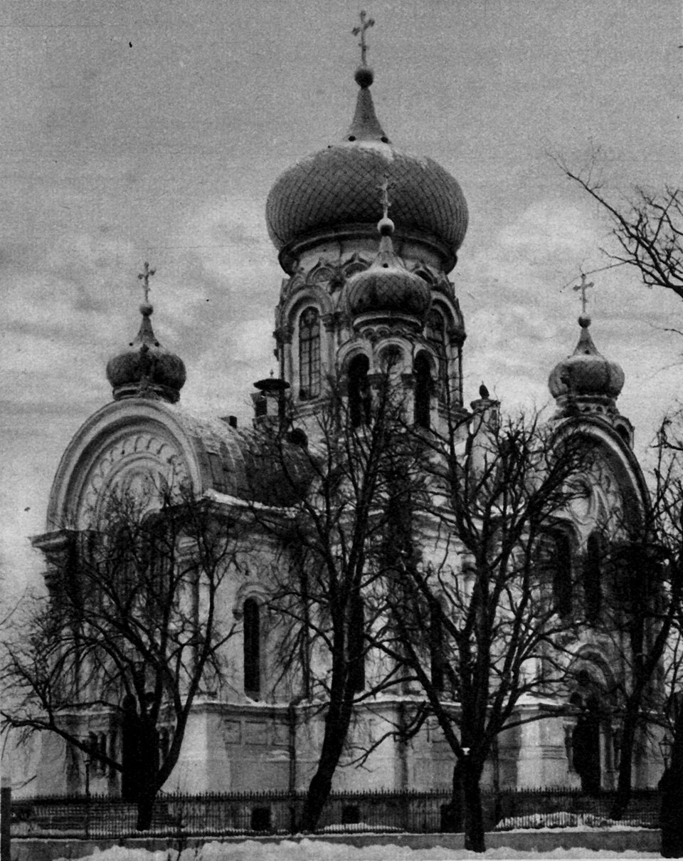 Fotografia przedstawia budynek z trzema wieżami zakończonymi kopułami na górze i krzyżem na czubkach stojący pośród drzew.
