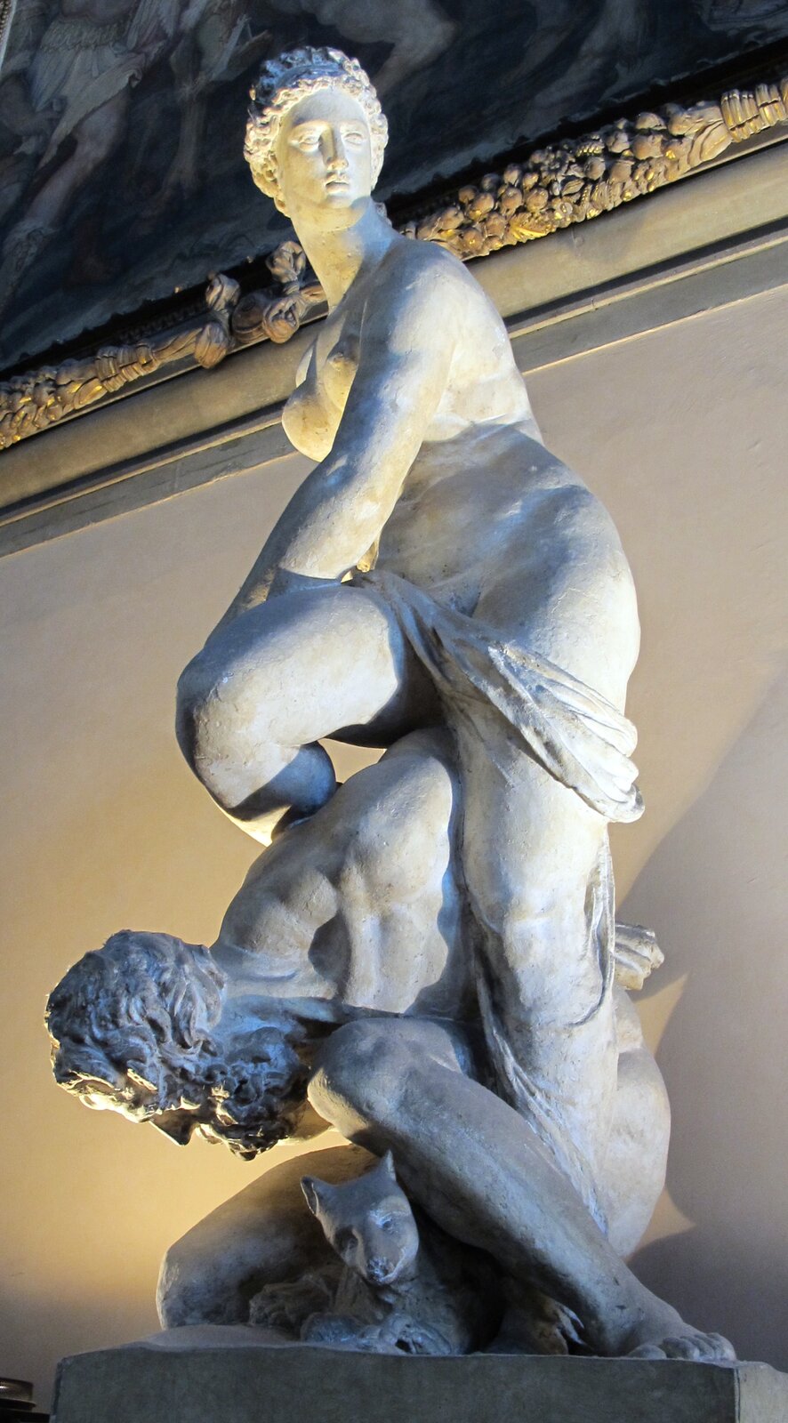 Wysoka, jasna rzeźba przedstawiająca nagą kobietę stojącą na plecach klęczącego mężczyzny; kobieta odwraca głowę w bok, a mężczyzna pochyla się w dół. U ich stóp widoczna jest głowa zwierzęcia przypominająca lwa. Całość ustawiona jest we wnętrzu, pod dekorowanym sufitem, oświetlona ciepłym światłem.