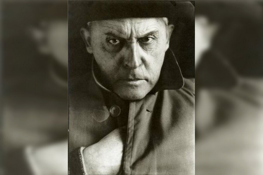 Czarno‑biała fotografia portretowa. Mężczyzna w średnim wieku patrzy prosto w obiektyw, z wyrazem intensywnej koncentracji. Ma głęboko osadzone oczy i zmarszczone brwi. Ubrany jest w płaszcz z podniesionym kołnierzem, lewą rękę trzyma wewnątrz płaszcza. Na głowie ma ciemne nakrycie.