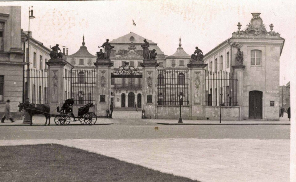 Ilustracja przedstawia Pałac Brühla w Warszawie. Ilustracja ukazuje widok szeroki, jasnego budynku z obszernym oknami. Przed pałacem jest wysokie ogrodzenie z szeroką, otwartą bramą wjazdową. Pomiędzy przęsłami ogrodzenia znajdują się ozdobne kolumny, na których są ustawione rzeźby. Przed pałacem widoczna jest ulica, którą jedzie dorożka.