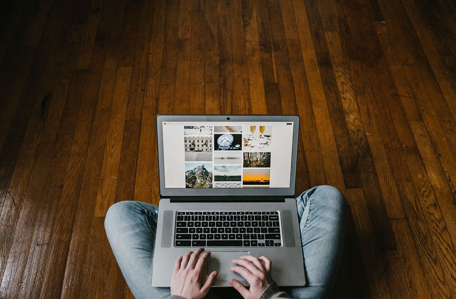  Zdjęcie przedstawia uda i ręce osoby siedzącej po turecku na drewnianej podłodze. Pomiędzy kolanami widoczny jest otwarty laptop, na ekranie którego widać różne zdjęcia, w tym: fasadę budynku, płonące ognisko, taflę wody, dwa kieliszki wypełnione płynem, górę i zegar. Obie dłonie spoczywają na laptopie.