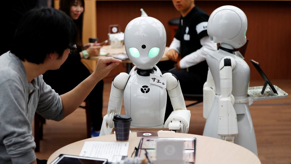 Na ilustracji widać fragment pomieszczenia, widać mężczyznę i dwa białe roboty. Jeden z nich stoi przy stole, mężczyzna siedzi obok stołu i dotyka robota.  