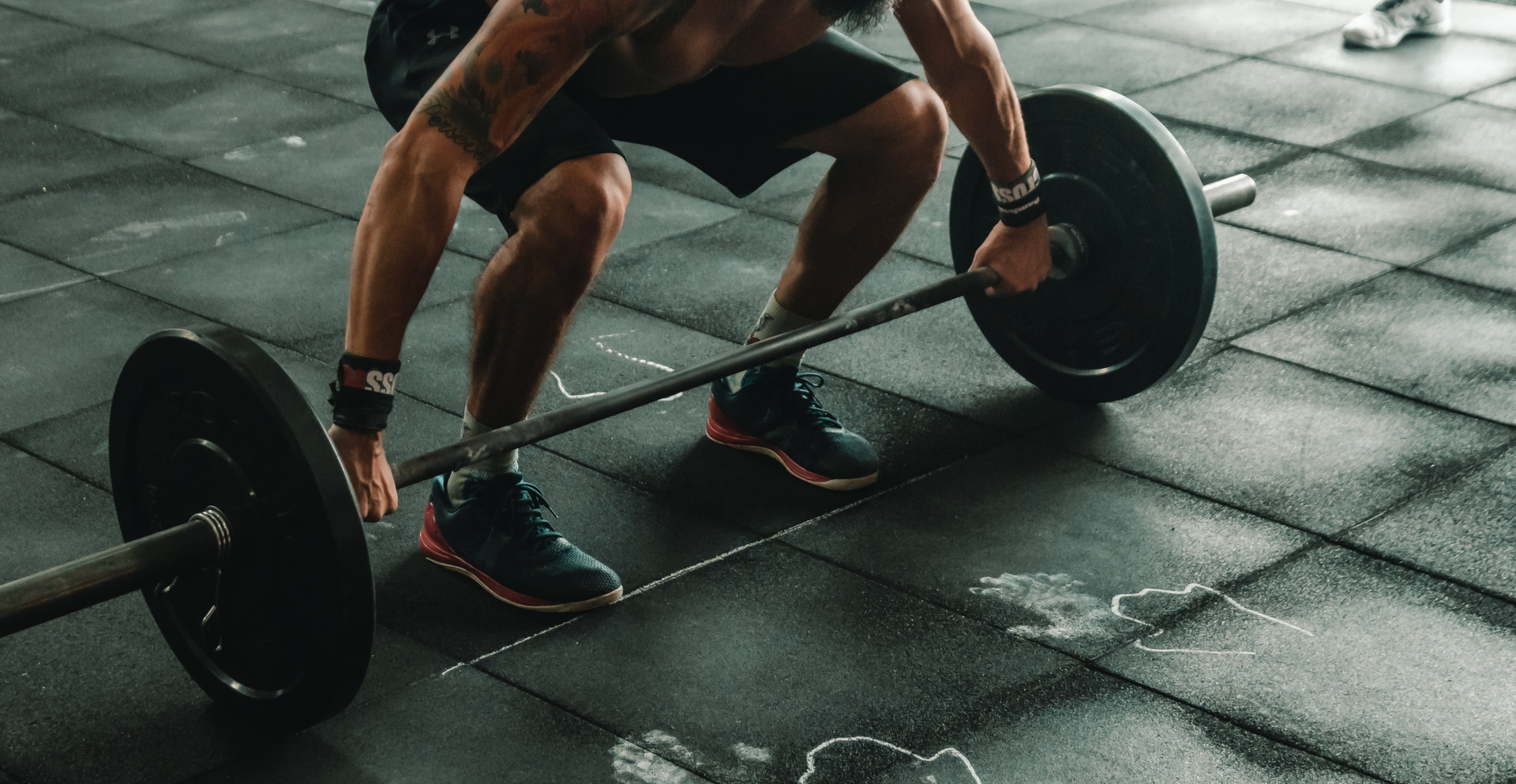 Zdjęcie przedstawia sylwetkę mężczyzny kucającego przed sztangą. Mężczyzna zamierza się do podniesienia ciężaru. Niewidoczna jest jego twarz.