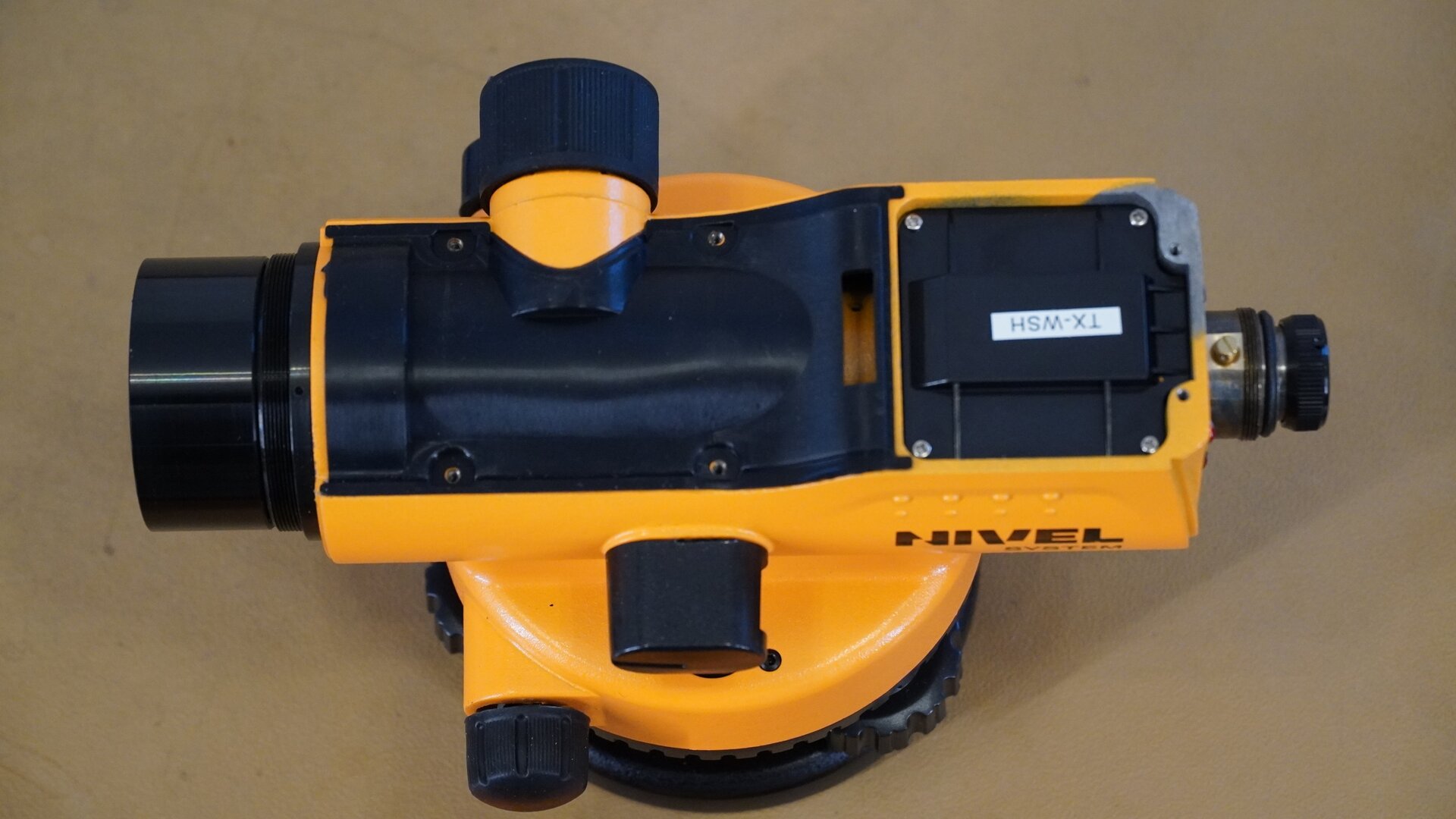 Na zdjęciu przedstawiono widok niwelatora z góry po demontażu osłony. Widoczne są śruby mocujące pokrywę oraz zespół okularu z krzyżem nitek. Pierścień okularu ma postać czarnego pokrętła, które reguluje ostrość krzyża nitek. Tuż za nim zabudowany jest kompensator.