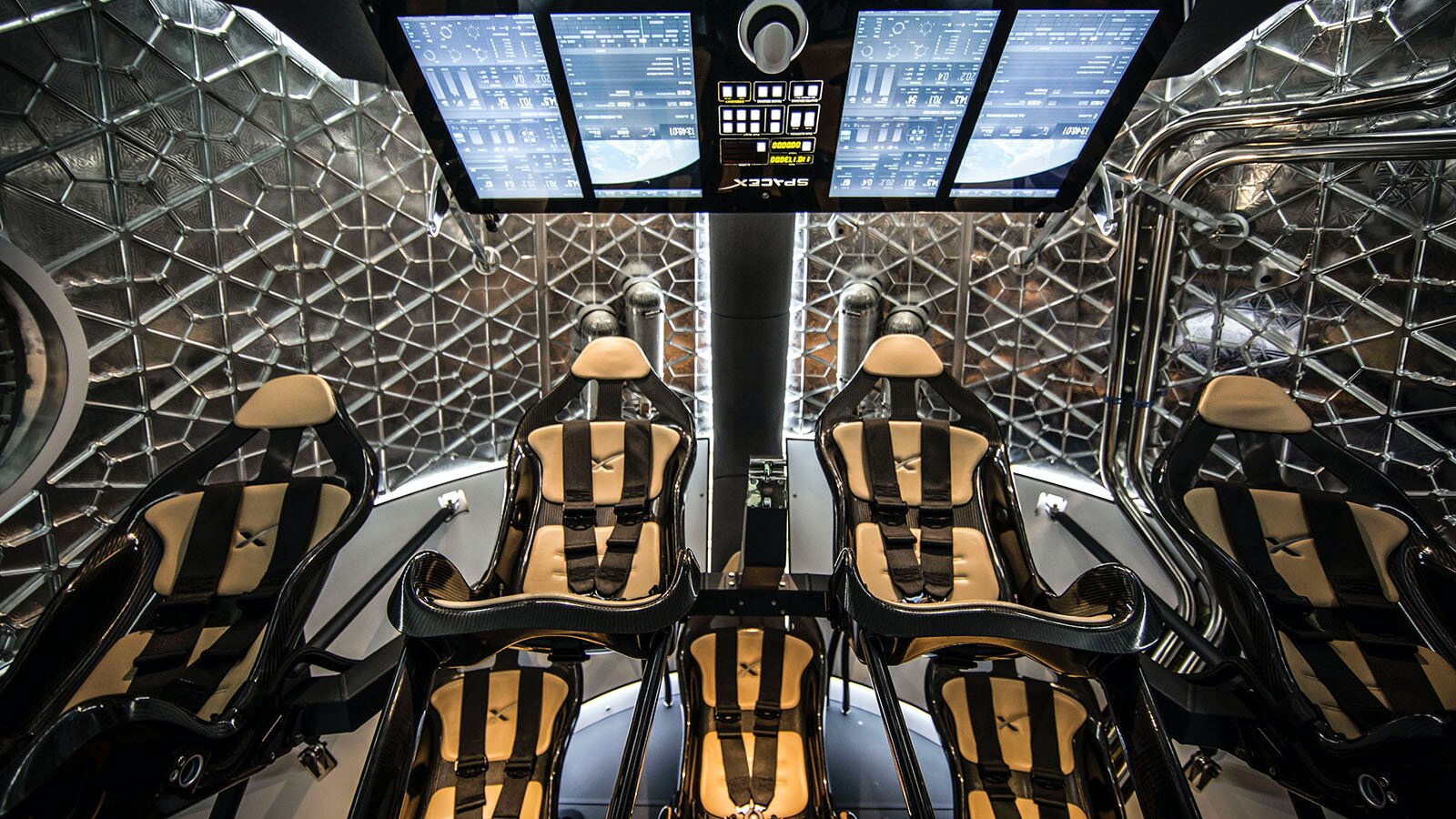 Zdjęcie przedstawia wnętrze kabiny statku kosmicznego. Na pierwszym planie widoczne są cztery fotele z pasami bezpieczeństwa. Powyżej widoczny jest monitor z czterema ekranami oraz panelem sterowania pośrodku. W tle widoczna jest metalowa, ażurowa konstrukcja przypominająca plaster miodu.