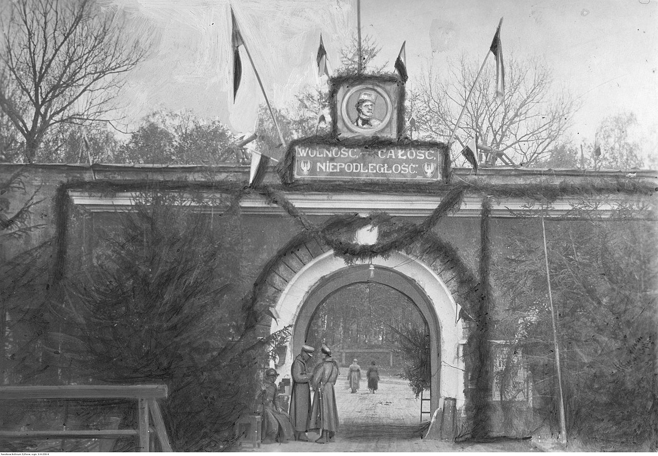 Kliknij, aby powiększyć. Ilustracja przedstawia bramę wejściową do twierdzy w Bobrujsku. Przy bramie znajduje się trzech wartowników, którzy bronią do niej dostępu. Wszyscy są w mundurach wojskowych. Jeden z nich siedzi, trzymając karabin z bagnetem oparty kolbą o ziemię. Przez otwartą bramę widać dwie osoby wchodzące do wnętrza twierdzy. Nad bramą znajduje się duży portret Tadeusza Kościuszki, a pod nim umieszczono napis: Wolność, Całość, Niepodległość. Ponad bramą umieszczone zostało sześć polskich sztandarów w barwach narodowych.