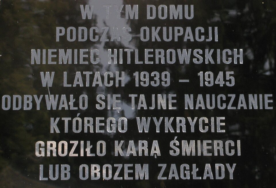 Ilustracja przedstawia tablicę upamiętniającą tajne nauczanie. Tablica wykonana jest z czarnego kamienia, napis na niej wygrawerowany jest w jasnym kolorze. Napis na tablicy: W tym domu podczas okupacji Niemiec hitlerowskich w latach 1939–1945 odbywało się tajne nauczanie, którego wykrycie groziło karą śmierci lub obozem zagłady. 
