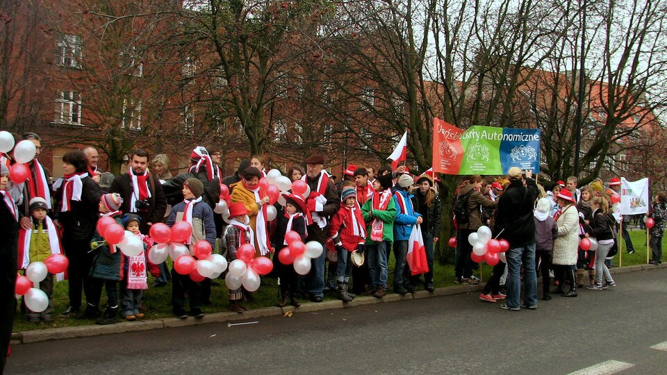 Zdjęcie przedstawia miejska ulicę. Przy krawężniku znajduje się duża grupa ludzi w biało‑czerwonych szalikach. Są też dzieci trzymające białe i czerwone balony
