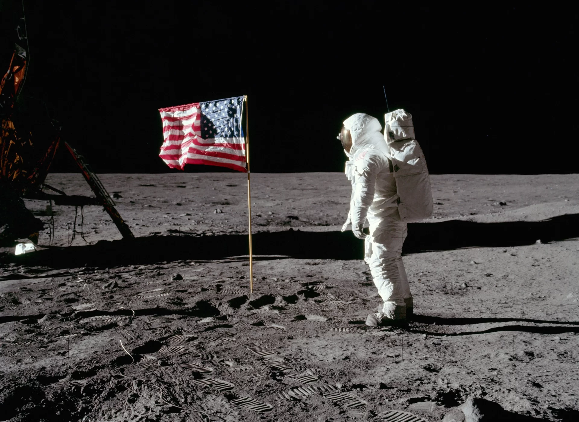 Na zdjęciu znajduje się człowiek ubrany w biały strój astronauty. Na plecach ma duży plecak, a jego głowę chroni duży hełm z szybą. Stoi on na księżycu koło kosmicznego pojazdu i wbitej w powierzchnię księżyca amerykańskiej flagi.
