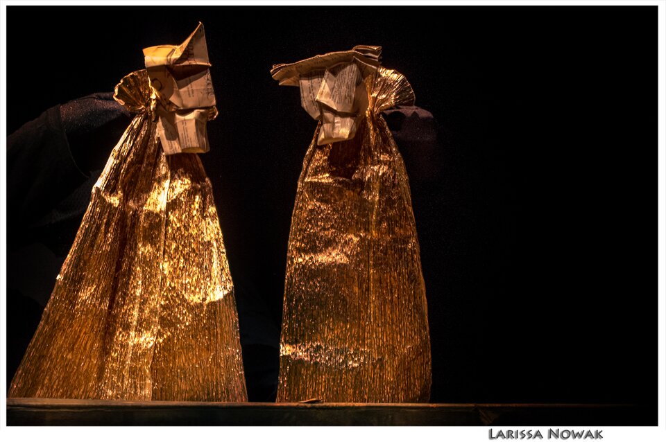 Ilustracja przedstawia dłonie w czarnych rękawiczkach trzymające postacie stworzone ze złotego papieru. Postacie są schematyczne, to paski papieru rozszerzone ku dołowi, z zebranym na górze końcem, na którym doczepione są papierowe elementy imitujące głowy.