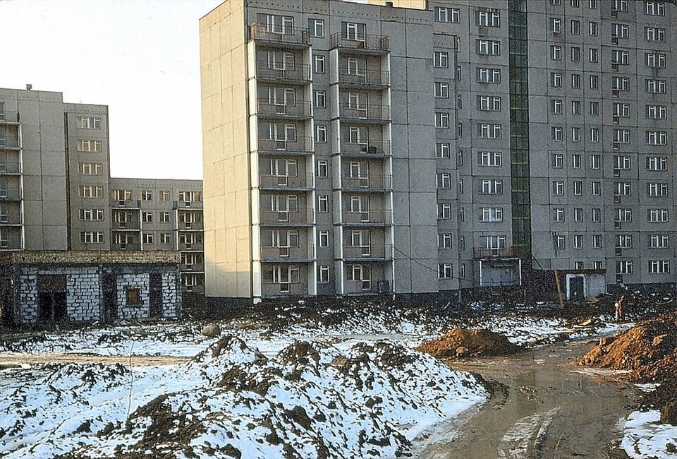 Fotografia przedstawia kilka bloków mieszkalnych zbudowanych z tak zwanej wielkiej płyty. Wszystkie są do siebie podobne: mają okna i balkony. Na pierwszym planie znajduje się droga wyjeżdżona przez samochody. Prowadzi ona w stronę bloków. Przy drodze znajdują się pojedyncze kopce ziemi pokryte śniegiem.