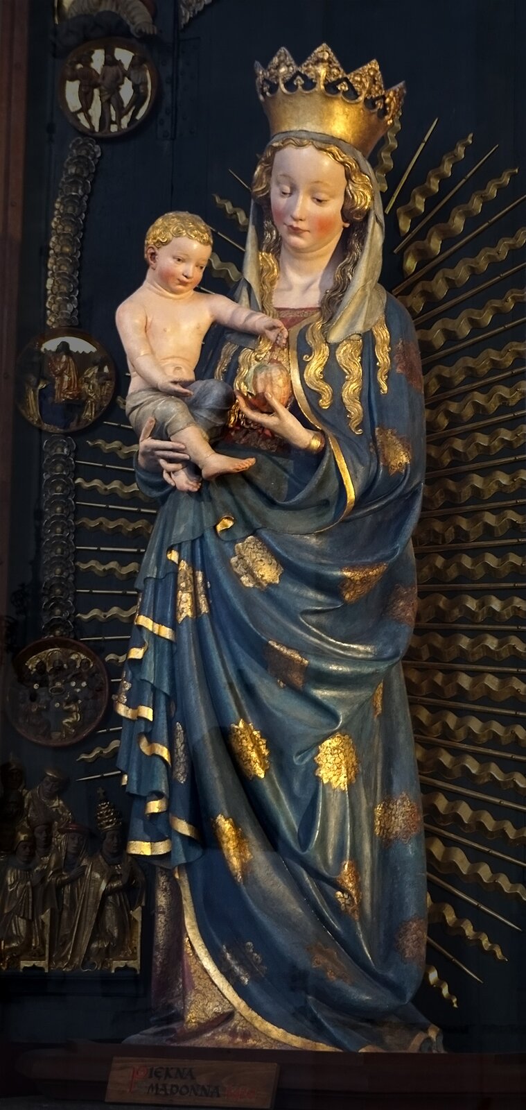 Ilustracja przedstawia figurę Madonny trzymającej dziecko na prawej ręce. Madonna ubrana jest w niebieską szatę ze złotymi dodatkami oraz na głowie ma koronę. Dziecko, które jest przez nią trzymane, ma narzuconą szatę zakrywającą dolną część ciała. Kobieta jak i dziecko mają jasne włosy.