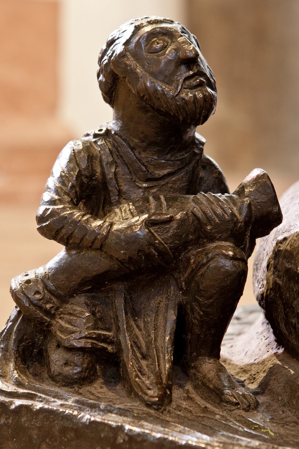 Detal romańskiej rzeźby z brązu: siedząca postać mężczyzny o brodatej twarzy, trzymająca na kolanach duży zwój lub księgę; powierzchnia rzeźby jest masywna i uproszczona, z wyraźnymi fałdami szat, tło rozmyte.